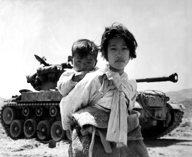 A frightened Korean girl carrying a baby on her back. Tanks can be seen in the background.